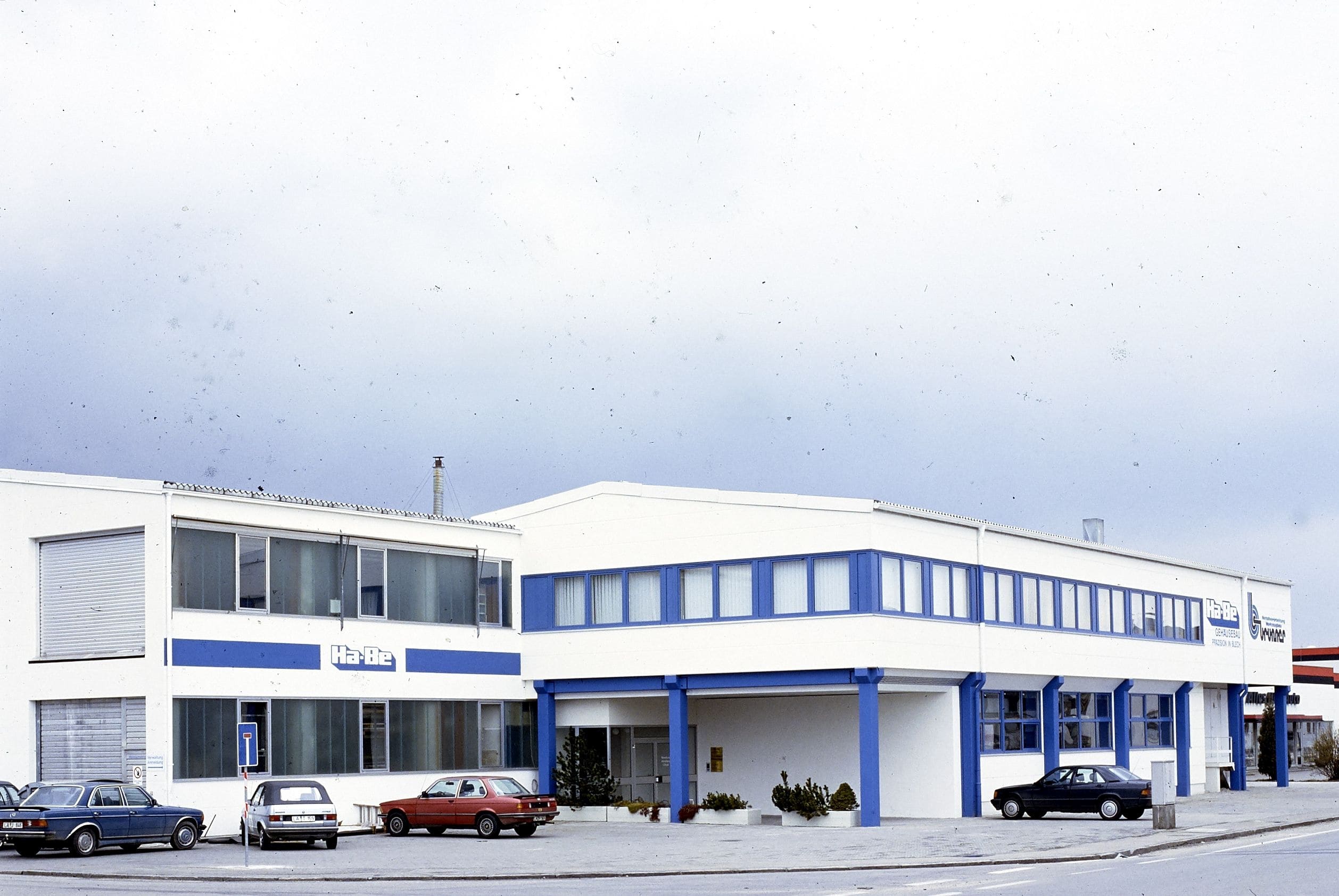 Ein weißes Geschäftsgebäude mit blauen Akzenten und großen Fenstern, vor dem an einem bewölkten Tag mehrere Autos geparkt sind. Das Gebäude hat Schilder und zwei Haupteingänge.