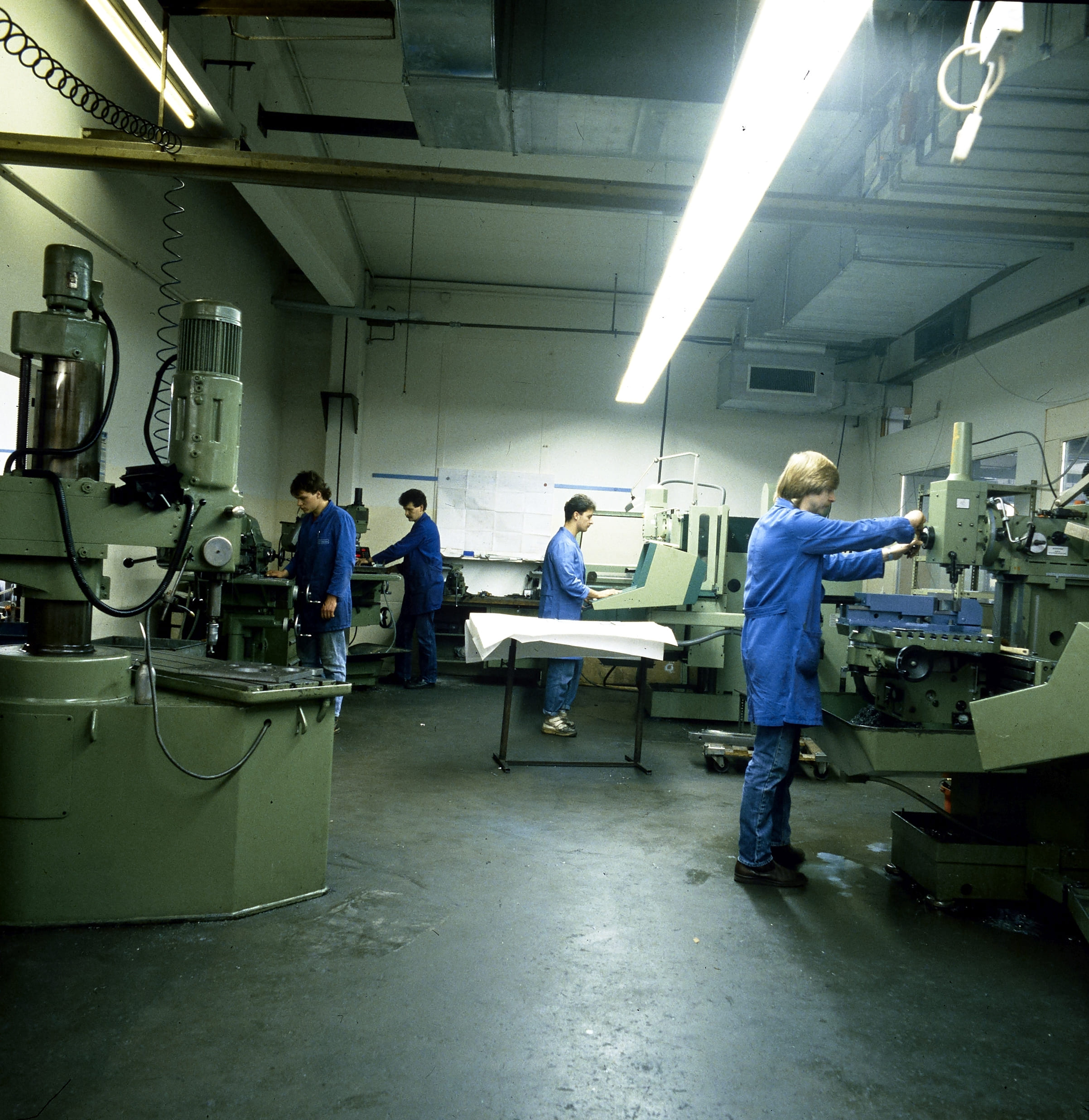 Mehrere Personen in blauen Laborkitteln bedienen große Industriemaschinen in einer geräumigen, gut beleuchteten Fabrik oder Werkstatt mit Betonböden und Leuchtstoffröhren über dem Kopf.