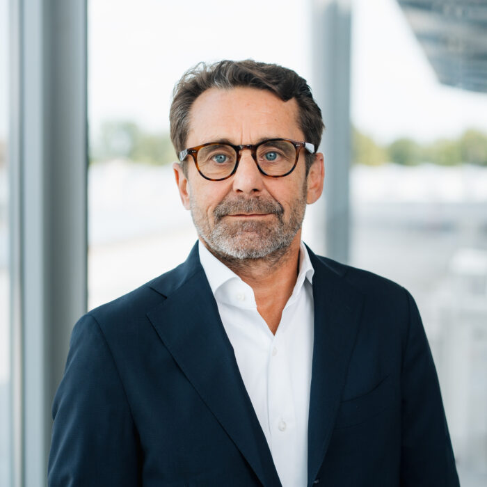 Ein Mann mittleren Alters mit kurzen braunen Haaren, Bart und Brille trägt einen dunklen Anzug und ein weißes Hemd und steht in einem Haus in der Nähe großer Fenster mit einem unscharfen Hintergrund.
