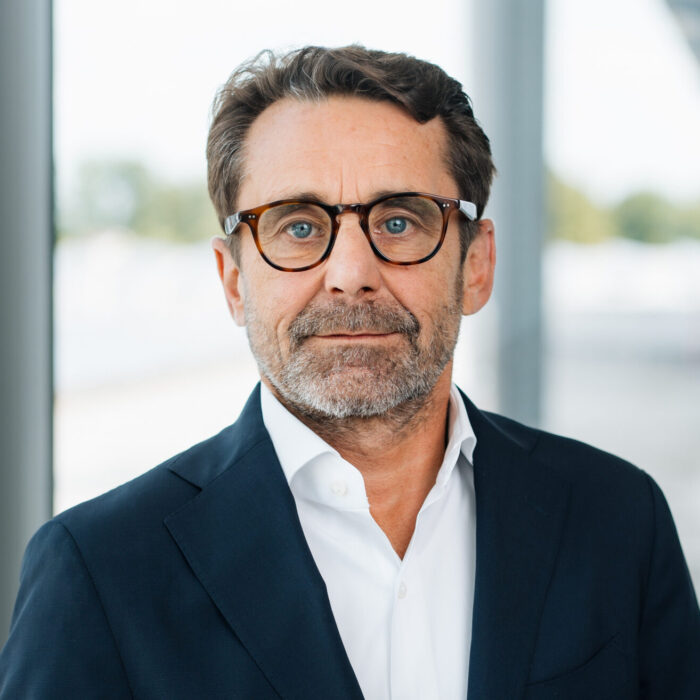 Ein Mann mittleren Alters mit kurzen braunen Haaren, Bart und Brille trägt einen dunklen Anzug und ein weißes Hemd und steht in einem Haus in der Nähe großer Fenster mit einem unscharfen Hintergrund.