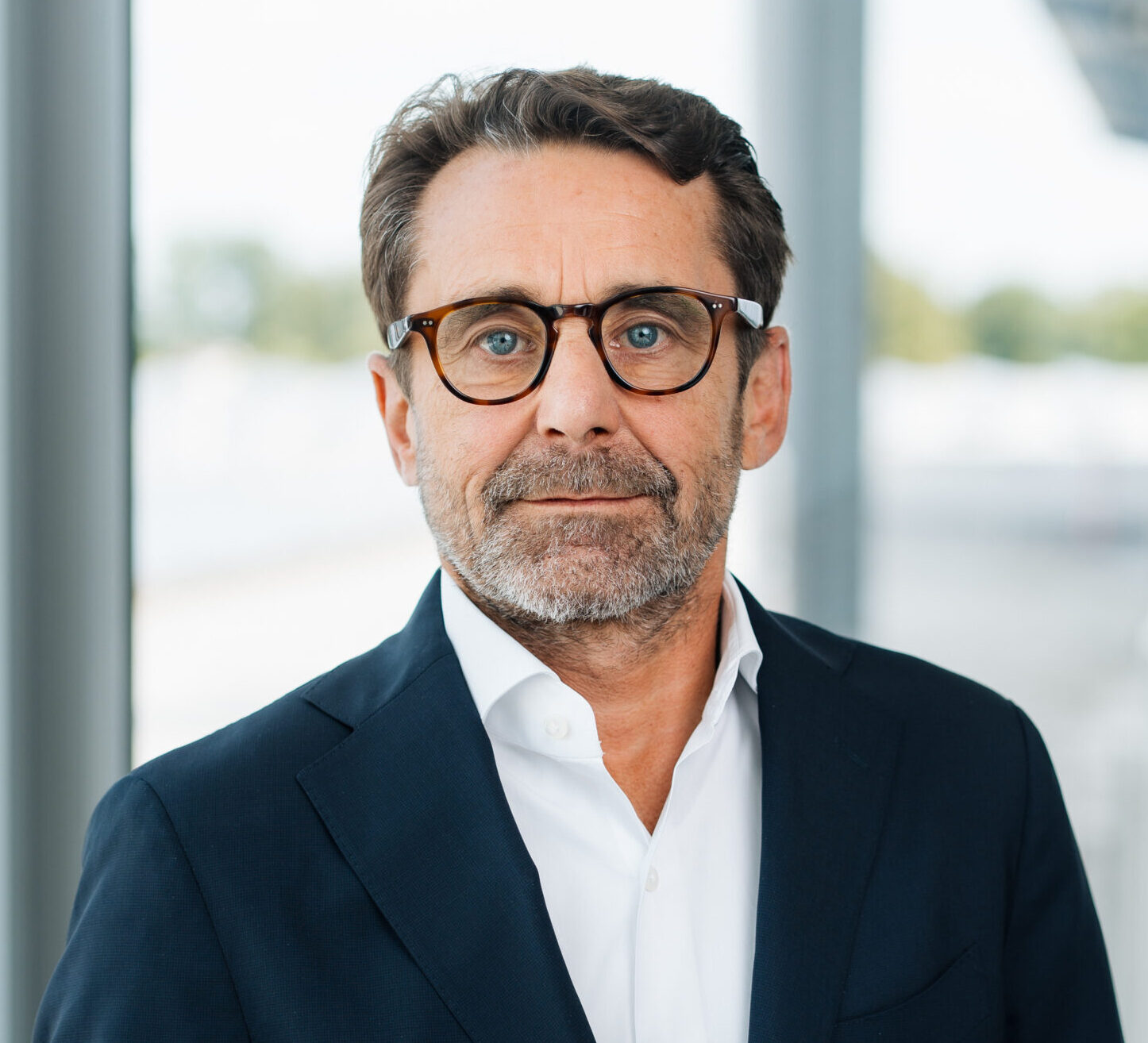 Karl-Heinz Brunner Ein Mann mittleren Alters mit kurzen braunen Haaren, Bart und Brille trägt einen dunklen Anzug und ein weißes Hemd und steht in einem Haus in der Nähe großer Fenster mit einem unscharfen Hintergrund.