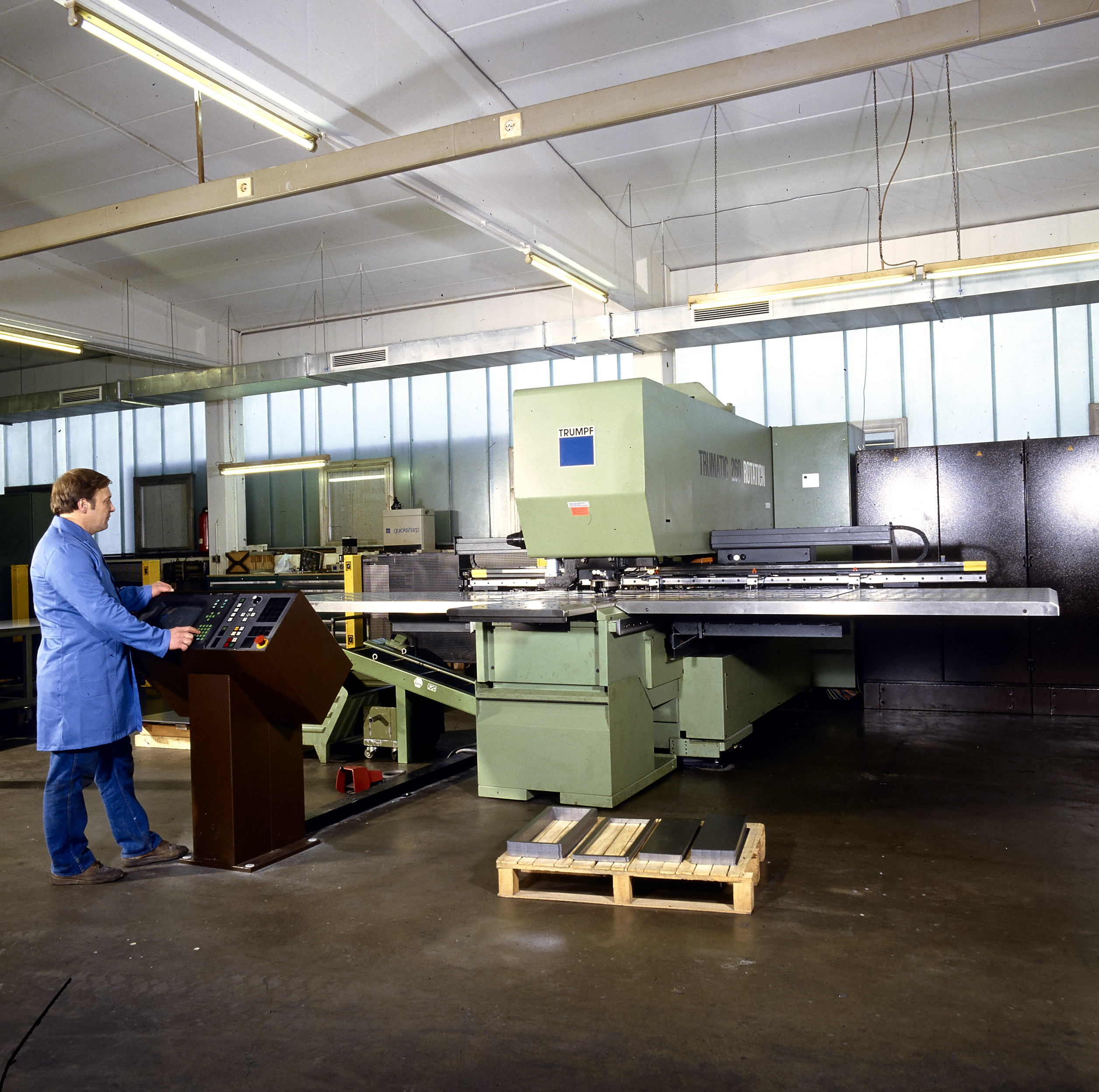 Ein Mann in einem blauen Laborkittel bedient eine große grüne Industriemaschine mit einem Schaltpult in einer geräumigen Fabrikhalle. Im Hintergrund sind Paletten auf dem Boden und Metallkonstruktionen zu sehen.