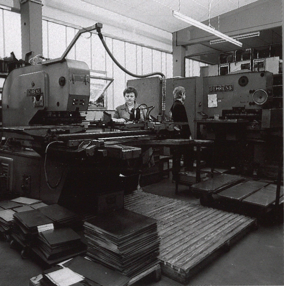 Zwei Personen arbeiten in einer Fabrik mit großen Behrens-Maschinen, umgeben von Blechstapeln und Geräten, in einem Raum mit großen Fenstern und Oberlichtern.