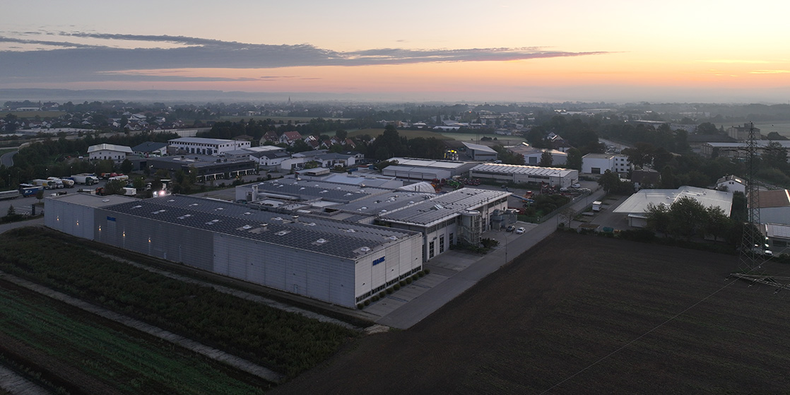 Luftaufnahme eines Industriekomplexes mit großen Gebäuden und Sonnenkollektoren auf den Dächern, umgeben von Feldern und Wohnhäusern bei Sonnenaufgang. Der Himmel ist teilweise bewölkt mit weichem, frühmorgendlichem Licht.
