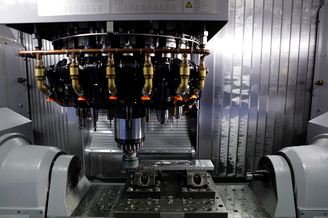 Nahaufnahme einer CNC-Fräsmaschine in Betrieb, mit einer rotierenden Spindel und verschiedenen Werkzeugköpfen, die über einem Metallwerkstück in einer Industrieumgebung positioniert sind.