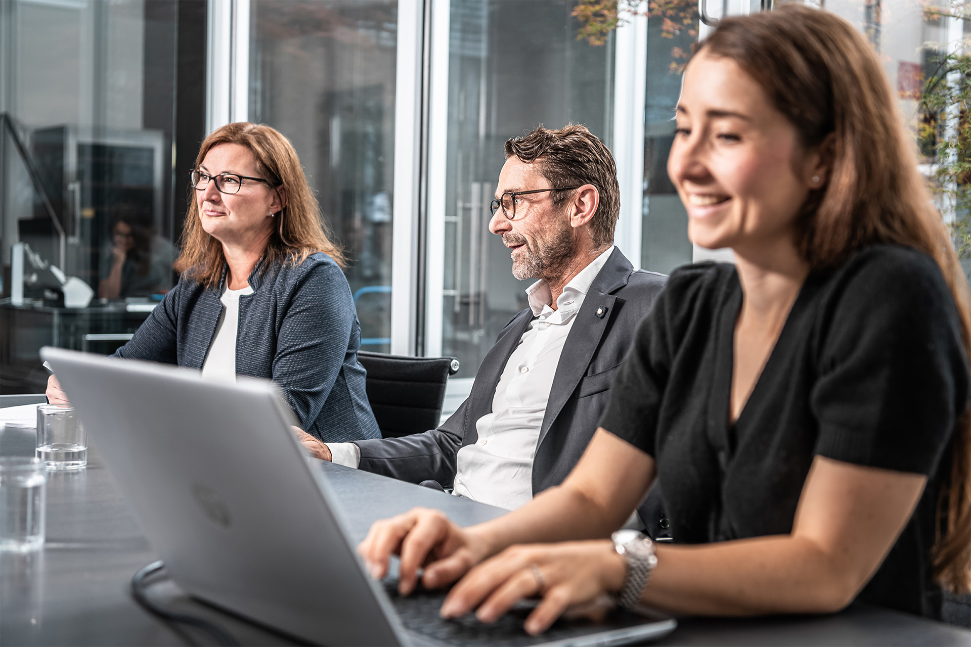 Drei Personen sitzen an einem Konferenztisch in einem modernen Büro. Zwei hören aufmerksam zu, einer lächelt und tippt auf einem Laptop. Große Fenster im Hintergrund lassen natürliches Licht herein.