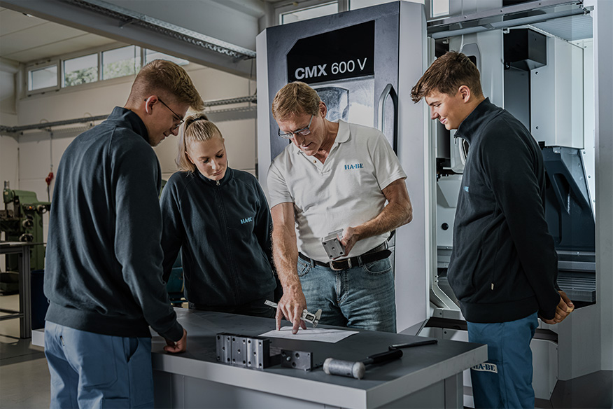 Ein Mann in einem weißen Polohemd erklärt drei jungen Erwachsenen in dunklen Jacken, die in einer Industriehalle um einen Tisch mit technischen Zeichnungen und Maschinenteilen versammelt sind, ein Metallteil.