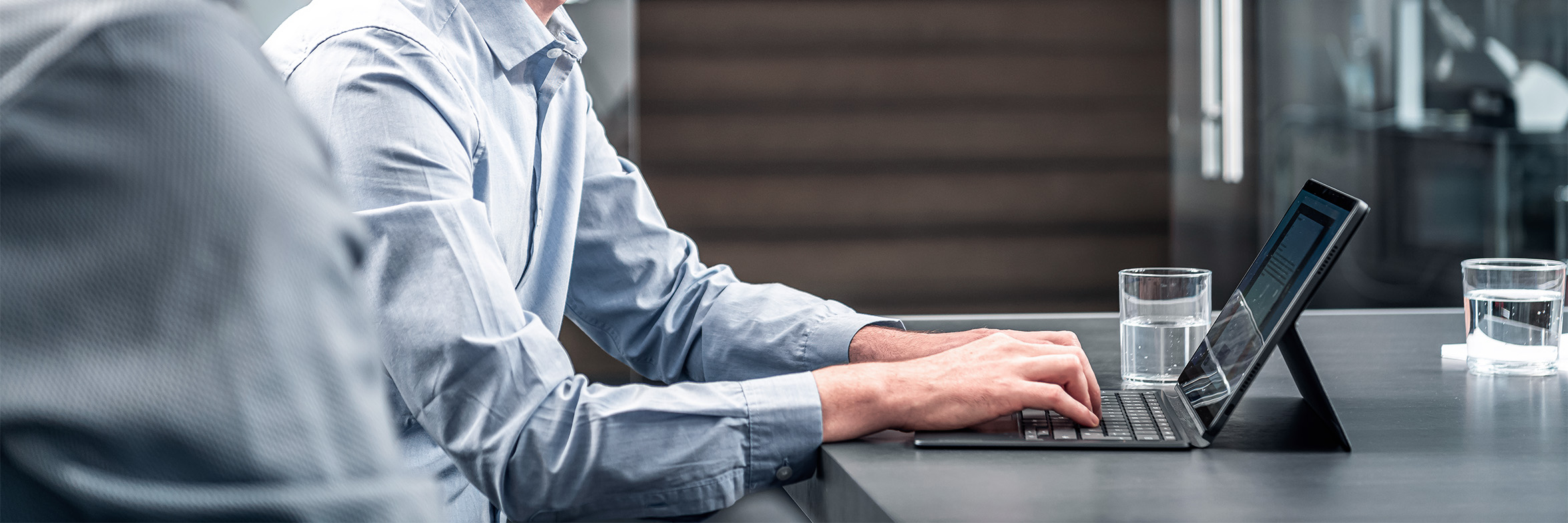 Zwei Personen in Geschäftskleidung sitzen an einem Tisch mit Wassergläsern; einer tippt auf einem Laptop, während der andere teilweise sichtbar ist, was auf eine professionelle Besprechung oder Zusammenarbeit in einer modernen Büroumgebung hindeutet.