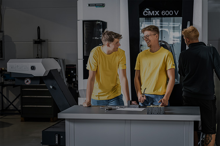 Zwei junge Männer in gelben Hemden stehen in einer modernen Industriehalle und lächeln sich an, während eine weitere Person in der Nähe steht und den Rücken zudreht. Im Hintergrund sind Maschinen und Arbeitsgeräte zu sehen.