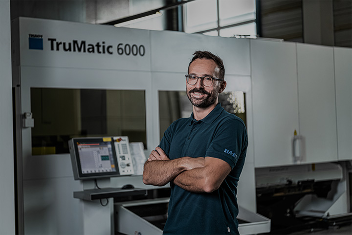 Ein Mann mit Brille und Bart, der ein dunkles Poloshirt trägt, steht mit verschränkten Armen und lächelt vor einer großen TruMatic 6000-Industriemaschine in einer modernen Fabrikumgebung.