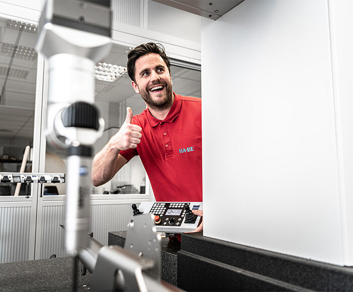 Ein Mann in einem roten Polohemd lächelt und zeigt den Daumen nach oben, während er ein Bedienfeld in einer hellen, modernen Labor- oder Fabrikumgebung mit technischen Geräten im Vordergrund hält.