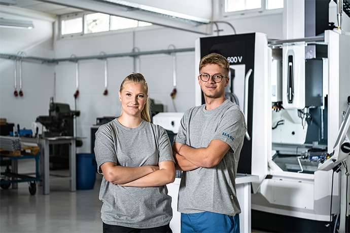 Zwei junge Erwachsene, eine Frau und ein Mann, stehen mit verschränkten Armen nebeneinander und lächeln in einer modernen Industriehalle mit großen Maschinen und heller Beleuchtung im Hintergrund.