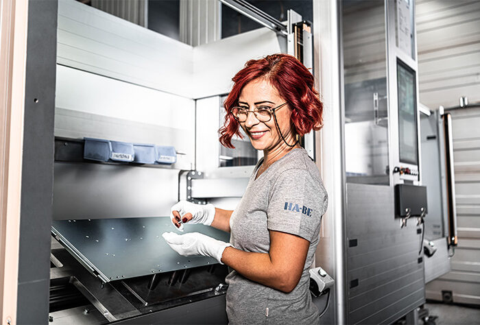 Eine Frau mit roten Haaren und Brille, die weiße Handschuhe und ein graues Hemd trägt, steht lächelnd an einer Maschine oder Ausrüstung in einer modernen Industrieumgebung.