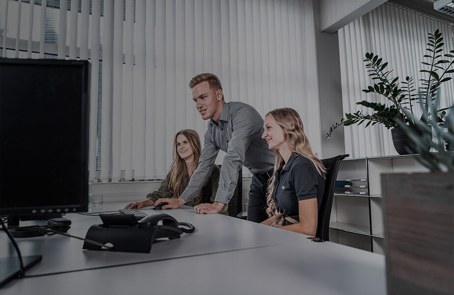 Drei Personen arbeiten in einem Büro an einem Schreibtisch mit Computern. Ein Mann steht, während zwei Frauen sitzen, alle auf den Computerbildschirm konzentriert. Der Raum hat große Fenster mit Jalousien und moderne Büromöbel.