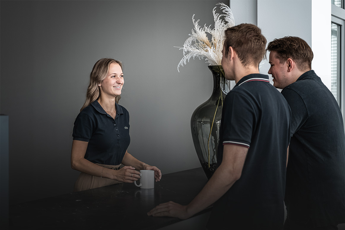 Eine Frau steht hinter einem dunklen Tresen, lächelt und hält eine Tasse in der Hand, während sie sich mit zwei Männern unterhält, die auf der anderen Seite stehen. Auf dem Tresen steht eine große Vase mit Ziergräsern.