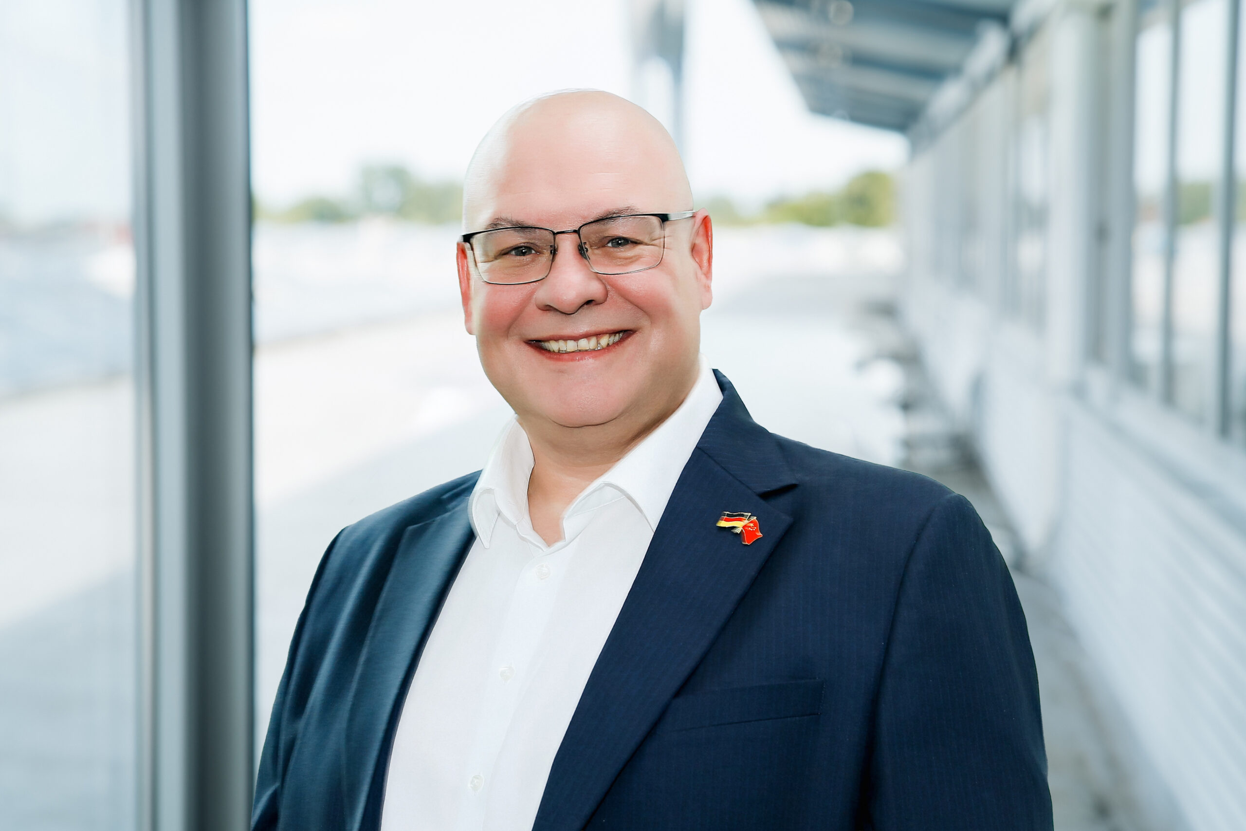 Ein Mann in blauem Blazer und weißem Hemd steht lächelnd in einem hellen, modernen Korridor mit großen Fenstern und weißen Wänden.