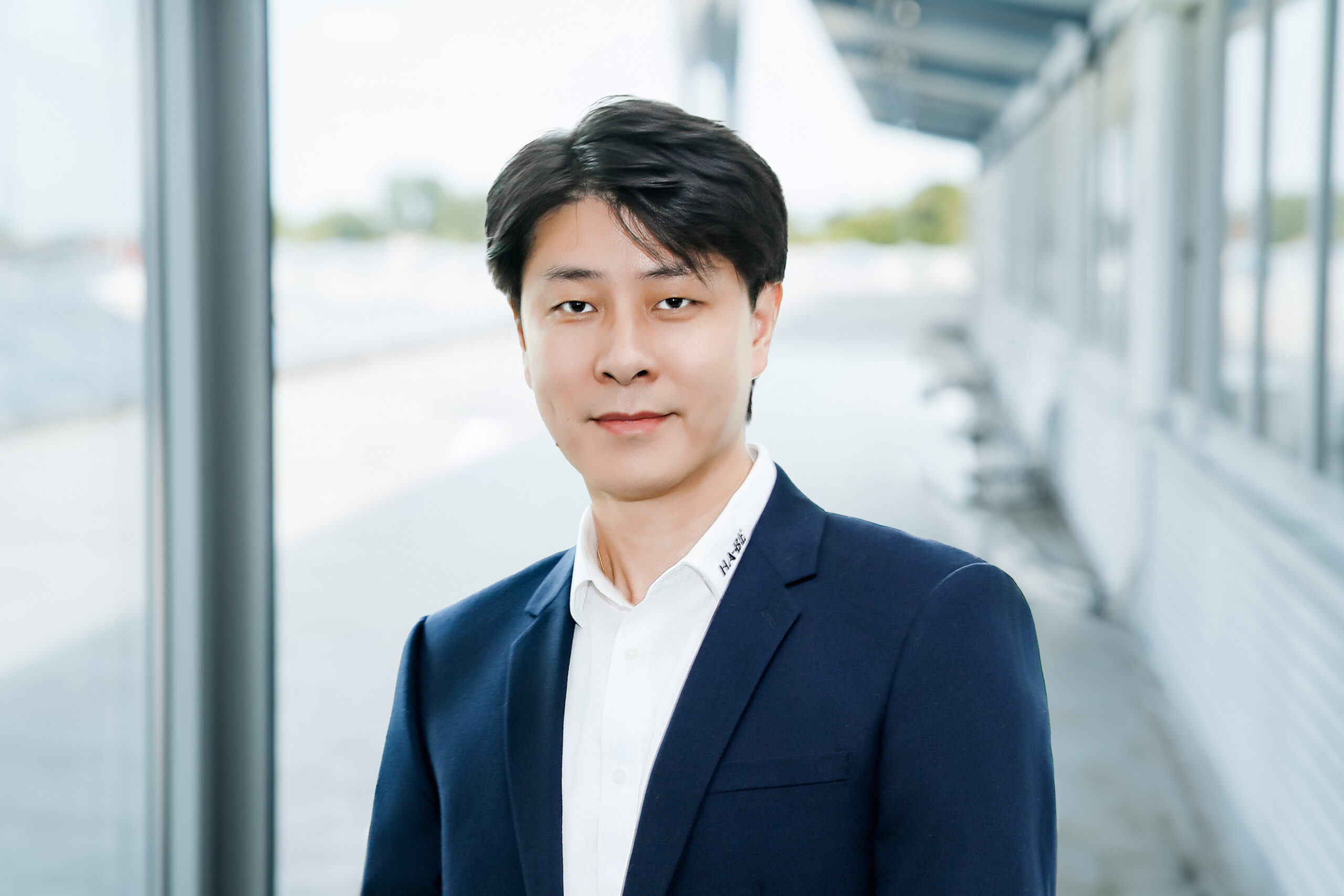 Ein Mann in einem marineblauen Anzug lächelt sanft in die Kamera. Im Hintergrund sind große Fenster und eine helle, moderne Umgebung mit verschwommenen Details zu sehen.