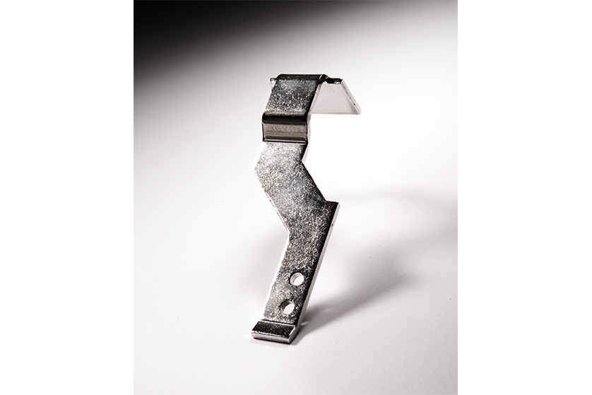Ein metallischer, zickzackförmiger Bügel mit zwei Löchern an einem Ende, der aufrecht auf einer weißen Fläche steht und hinter dem sich ein Schatten befindet.