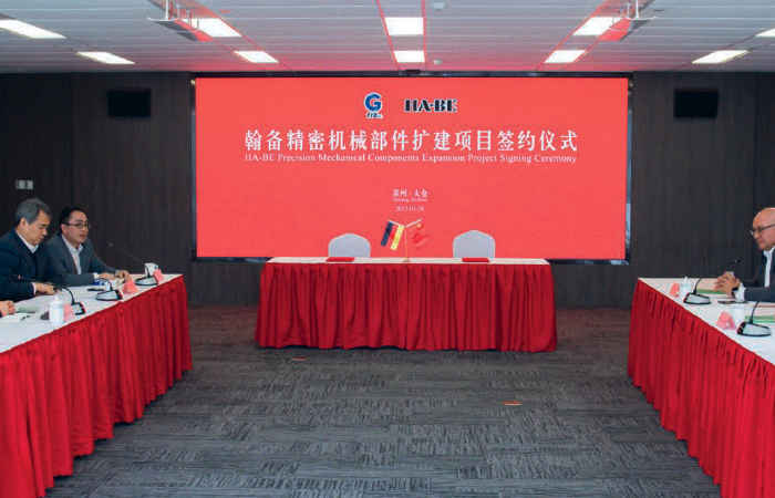 Zwei Gruppen von Personen in Geschäftskleidung sitzen sich in einem Konferenzraum an Tischen gegenüber. Ein großer roter Bildschirm hinter ihnen zeigt chinesischen Text und die Worte 