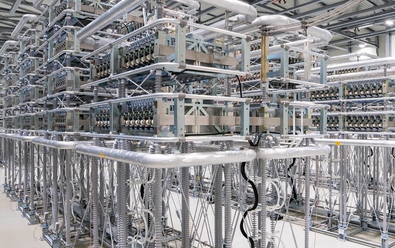 Großer Industrieraum voller komplexer Metallstrukturen, Kabel und elektronischer Komponenten, der wahrscheinlich ein Umspannwerk für das Stromnetz oder ein Datenzentrum mit fortschrittlicher Elektro- und Computerausrüstung darstellt.