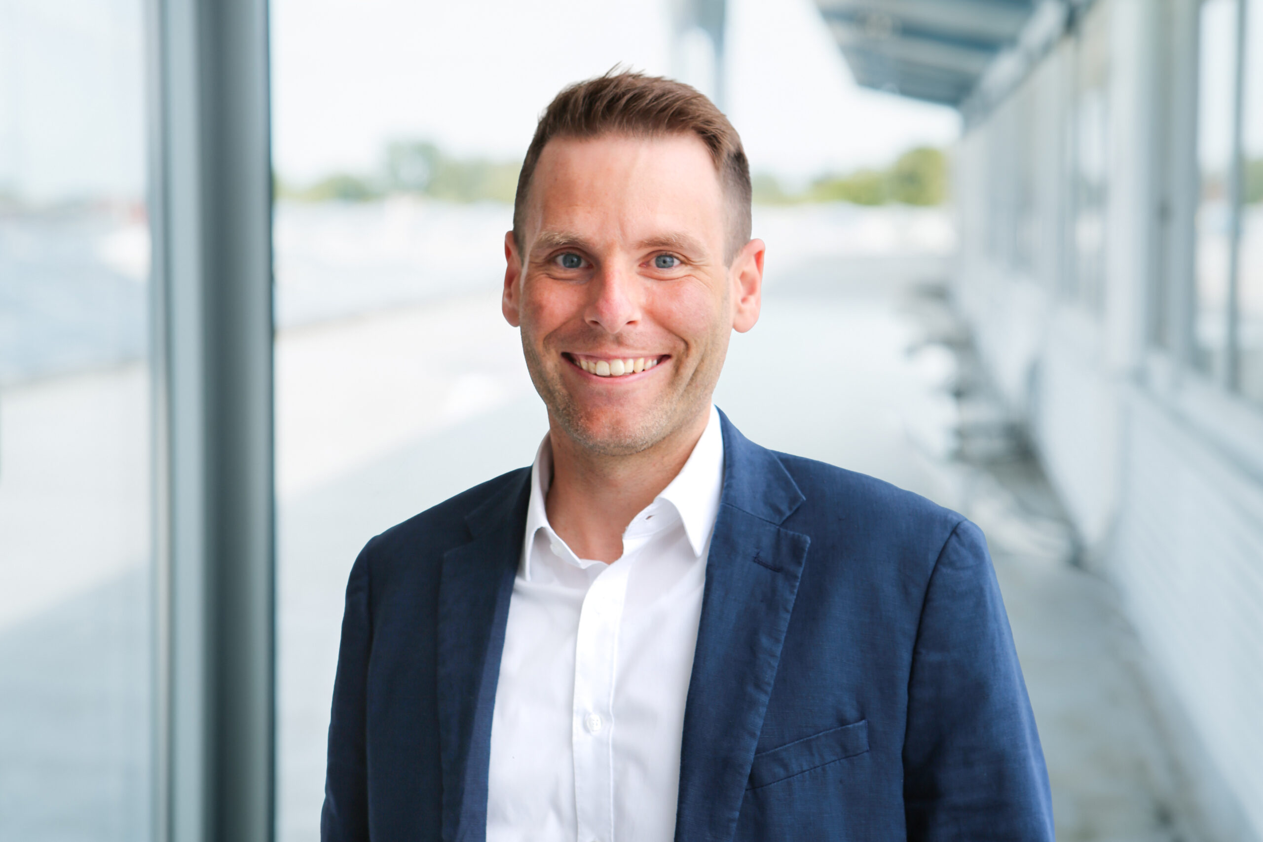 Ein Mann in blauem Blazer und weißem Hemd steht lächelnd in einem hellen, modernen Korridor mit großen Fenstern und weißen Wänden.