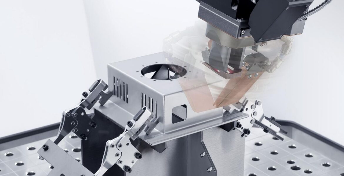 Ein Roboterarm mit einem Schweiß- oder Schneidewerkzeug arbeitet an einem rechteckigen Metallkasten, der mit Klemmen auf einer perforierten Metallarbeitsfläche in einer Hightech-Industrieumgebung befestigt ist.