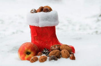 Ein roter, mit Nüssen gefüllter Weihnachtsstrumpf liegt im Schnee, umgeben von Walnüssen, einem roten und gelben Apfel und einem Tannenzapfen.
