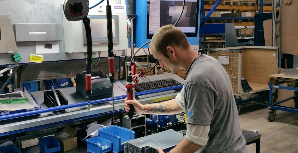 Ein Mann in einem grauen Hemd arbeitet an einem Arbeitsplatz in einer Fabrik mit einem Elektrowerkzeug an elektronischen Geräten. Auf der Werkbank und an der Wand hinter ihm sind ein großer Monitor und verschiedene Werkzeuge zu sehen.