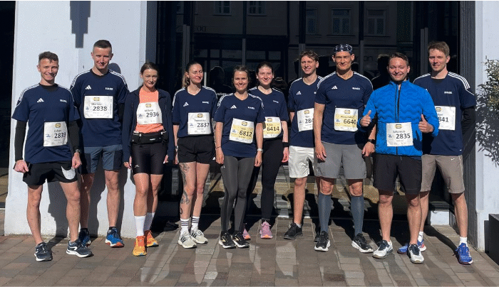 Eine Gruppe von zehn Läufern in Sportkleidung steht vor einem Rennen lächelnd beieinander, jeder trägt eine Startnummer. Das Wetter scheint sonnig und angenehm zu sein.